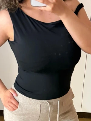 Svart oneshoulder topp - Snygg svart oneshoulder topp med tight passform. Toppen är ärmlös och har en asymmetrisk axelrem som ger en trendig look. Perfekt att styla med både jeans och kjol för en cool outfit.