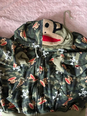 Paul Frank tröja - Cool camo hoodie från paul Frank dragkedja och huva. Hoodien har ett unikt apansikte broderat på huvan och camomönster med apor och dödskallar över hela plagget. Mjuka muddar och fickor framtill. Perfekt för streetwear-stilen.