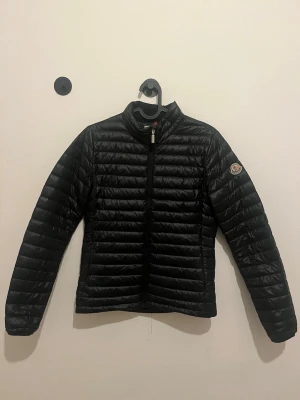 Svart dunjacka från Moncler - Snygg svart dunjacka från Moncler med quiltad design och hög krage. Perfekt nu till våren och riktigt lätt att bära. Klassisk och stilren modell som funkar till det mesta!