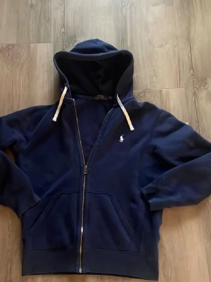 Mörkblå hoodie från Polo Ralph Lauren - Mörkblå hoodie med dragkedja från Polo Ralph Lauren. Klassisk design med vit broderad logga på bröstet, vita snören i huvan och två stora fickor framtill. Mjuk bomullskänsla och ribbade muddar vid ärmslut och nederkant.