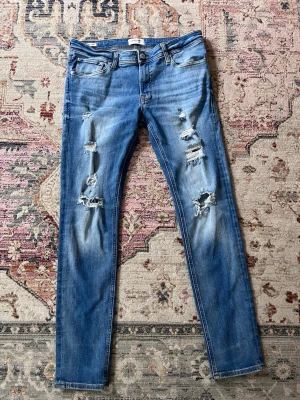 Blå slitna slimfit jeans från Jack&jones - Säljer ett par blå slimfit jeans från Jack&jones med slitningar och hål på framsidan. Jeansen har klassisk femficksmodell, låg midja och är tillverkade i stretchigt denim för en tajt passform. Perfekta för en avslappnad och trendig look.