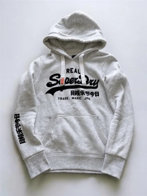 Grå hoodie från Superdry med tryck - Snygg ljusgrå hoodie från Superdry med svart tryck på bröstet och ärmen samt vita dragsnören i huvan. Klassisk känguruficka framtill och ribbade muddar. Perfekt för en avslappnad streetstyle-look. Skick 9/10, passar som xxs/xs