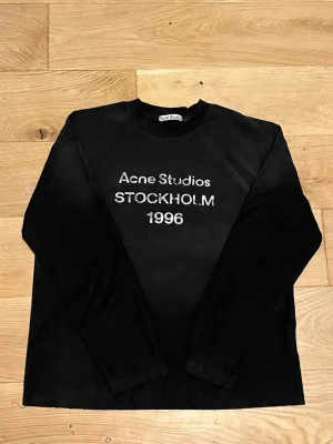 Svart långärmad topp Acne Studios - Svart långärmad topp från Acne Studios med vit broderad text 'Acne Studios STOCKHOLM 1996' på bröstet. Klassisk rund halsringning och rak passform. Perfekt för en stilren och trendig look.