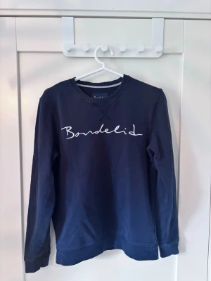 Mörkblå sweatshirt från Bondelid Storlek S - Mörkblå sweatshirt från Bondelid med vit broderad logga framtill. Klassisk rund halsringning och långa ärmar. Tillverkad i mjuk bomull.