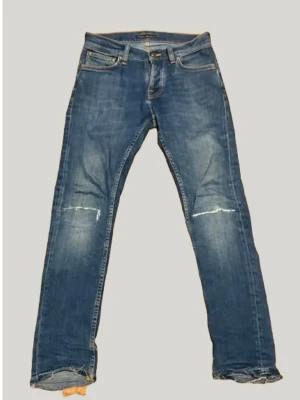 Blå raka jeans från Nudie Jeans - Snygga blå jeans från Nudie Jeans med passformen grim tim. Dem har slitna detaljer vid knäna och bensluten. Klassisk femficksmodell med och knapp. Perfekta för en avslappnad stil. Storlek 28/30