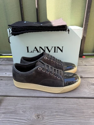 Lanvin gråa sneakers med lackad tå - Snygga gråa sneakers från Lanvin med ovandel i mocka och glansig lackad tå. Skorna har beige sula och klassisk låg profil med svarta skosnören och metallöljetter. Perfekta för dig som gillar stilrena och exklusiva detaljer. Dustbag och skor skickas med i beställningen. 