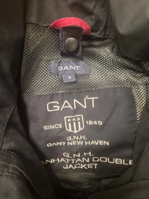 Svart vindjacka från GANT - Svart Gant Manhattan Double Jacket med dragkedja och knappar framtill. Jackan har en hög krage och en huva som kan fällas in. Det finns två fickor framtill med lock och knappar. Insidan av jackan är fodrad med ett nätmaterial och har två innerfickor med dragkedja i rött. Ärmsluten har knappar för justering.