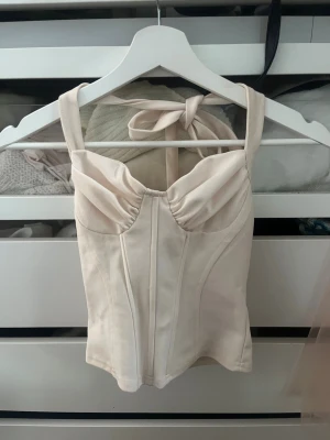 Beige korsettopp med knytband - Supersnygg beige korsettopp med breda axelband och knyt i nacken. Toppen har markerade sömmar framtill och en figurnära passform som ger en trendig siluett. Perfekt att styla med jeans eller kjol för en cool look.