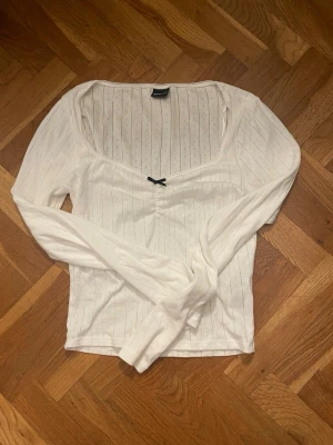 Vit långärmad topp med rosett - Supersöt vit långärmad topp från Gina Tricot med ribbat mönster och liten svart rosett framtill. Toppen har en v-ringad urringning. Aldrig använd. 