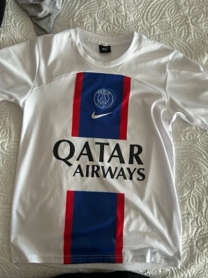 PSG Mbappé matchtröja Nike vit - Säljer en vit PSG matchtröja från Nike med blå och röda ränder framtill, Paris Saint-Germain-logga och Qatar Airways-tryck. På ryggen står det Mbappé och nummer 7. Perfekt för fotbollsfans som vill visa sitt stöd för laget.