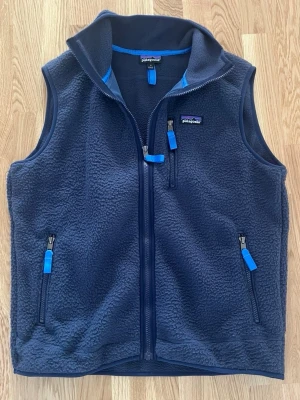 Mörkblå fleeceväst från Patagonia - Mysig mörkblå fleeceväst från Patagonia med tre fickor med dragkedja och blå detaljer. Västen har hög krage och är tillverkad i mjukt fleecematerial, perfekt för lager-på-lager och chill dagar.