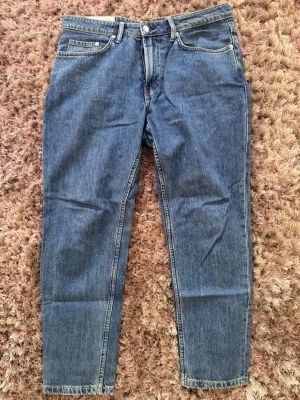 Blå raka jeans i klassisk modell - Säljer ett par blå jeans i klassisk rak modell med fem fickor och normal midja. Jeansen har en tidlös tvätt och är tillverkade i denim med synliga sömmar. Perfekta för en avslappnad och stilren look.