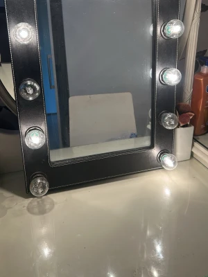 Svart sminkspegel med lampor - Stilren sminkspegel med svart ram och åtta stora LED-lampor runt spegeln för perfekt belysning. Rektangulär form och ram i ett material som ser ut som konstläder med vita sömmar. Perfekt för sminkning och styling vid sminkbordet.