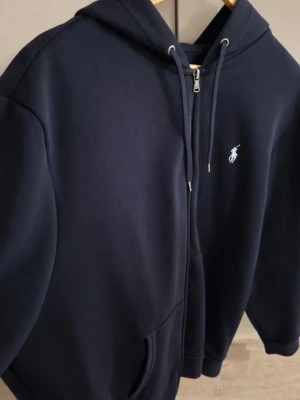 Svart zip-hoodie från Polo Ralph Lauren - Säljer en svart hoodie från Polo Ralph Lauren med dragkedja och huva. Klassisk design med logotyp broderad på bröstet, fickor framtill och snören i huvan. Materialet är dubbelstickad bomull som känns riktigt skön och premium.