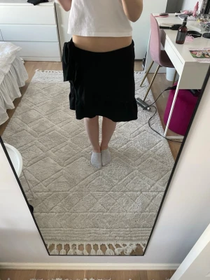 Svart omlottkjol från Vero Moda - Säljer en svart omlottkjol från Vero Moda i storlek S. Kjolen har en snygg knytning i sidan och är i en kort modell. Perfekt till sommaren och enkel att matcha med olika toppar.