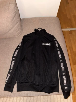 Svart zip-tröja från Hoodrich - Cool svart zip-tröja från Hoodrich med logga på bröstet och längs ärmarna. Tröjan har hög krage, ribbade muddar och dragkedja framtill. Perfekt för dig som gillar streetwear och vill ha en snygg och bekväm look.