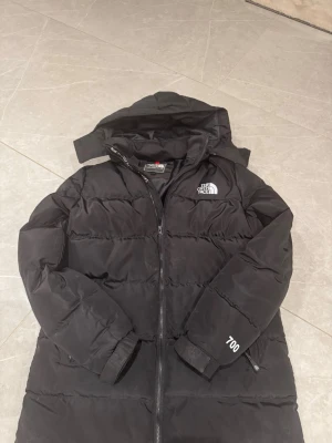 Svart långjacka från The North Face - Säljer en svart dunjacka från The North Face med huva och dragkedja framtill. Jackan har 700-fill power, broderad logga på bröstet och vänster ärm samt quiltad design. Perfekt för kalla vinterdagar och riktigt varm tack vare dunfyllningen. Storlek s!