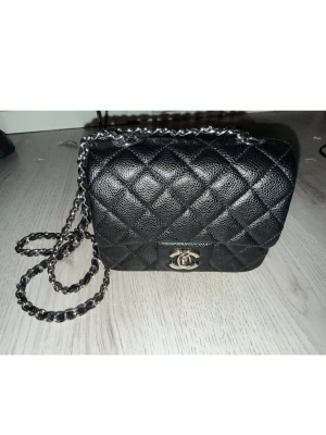 Svart Chanel Mini Square - Säljer en klassisk svart Chanel axelväska i quiltat läder med silverfärgad kedjerem och ikonisk CC-logga framtill. Väskan har lock och elegant design som ger en lyxig vibe. Perfekt för dig som vill ha en tidlös och trendig accessoar.
