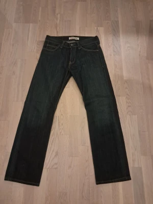 Mörkblå raka jeans från Levi's - Säljer ett par mörkblå jeans från Levi's med klassisk rak passform och fem fickor. Jeansen har kontrastsömmar och patch med Levi's-logga bak i midjan. Tillverkade i slitstarkt denimtyg, perfekta för en avslappnad och tidlös stil.