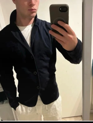 Mörkblå kavaj med klassisk passform - Säljer en stilren mörkblå kavaj med tre knappar och klassisk krage. Kavajen har raka ärmar och diskreta fickor framtill. Materialet ser ut att vara bomull eller en bomullsblandning, vilket ger en mjuk och bekväm känsla. Perfekt för dig som vill ha en enkel och snygg look.