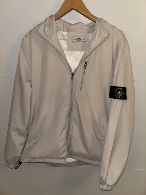 Beige vindjacka från Stone Island - Snygg beige vindjacka från Stone Island med ikonisk svart och gul patch på ärmen. Jackan har huva, dragkedja framtill, bröstficka med dragkedja och elastiska muddar. Tillverkad i lätt, vindtätt material som passar perfekt till streetwear.