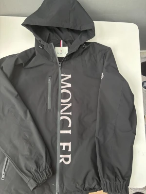 Svart vindjacka från Moncler - Stilren svart vindjacka från Moncler med stor logga tryckt vertikalt på framsidan. Jackan har huva, dragkedja framtill, bröstficka med dragkedja och elastiska muddar. Moncler-logga på ärmen och liten detalj i rött, vitt och blått på ryggen. Perfekt för en trendig streetwear-look.
