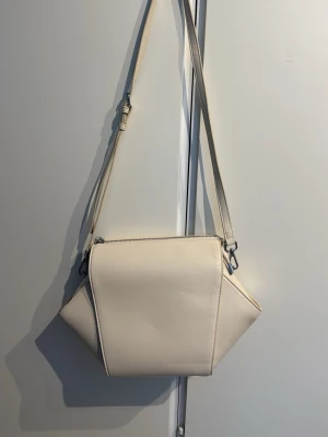 Krämvit axelväska - Säljer en krämvit väska som aldrig kommit till användning. Ljusterbart axelband, även avtagbart med rymligt innehåll med en liten ficka med dragkedja. 