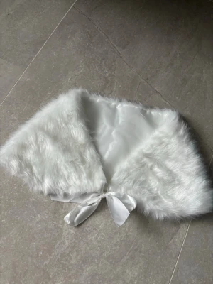 Vit fuskpäls sjal med sidenband - Superfin vit sjal i fuskpäls med glansigt sidenband att knyta framtill. Perfekt accessoar för att lyfta din outfit och ge en lyxig känsla. Mjuk och fluffig med slät insida i satinliknande material.