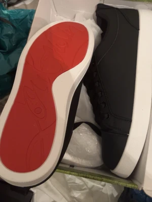 Svarta sneakers med röd sula - Snygga svarta sneakers från Christian Louboutin med klassisk röd sula och vita kanter. Skorna har en stilren design med svarta snören och diskreta sömmar. Perfekta för dig som vill ha något trendigt och exklusivt. Lite smutsiga bara vid sulan men kan fixa rent 