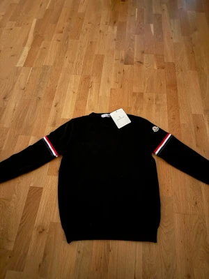Svart stickad tröja från Moncler - Svart stickad tröja från Moncler med rund halsringning och långa ärmar. Ärmarna har röd, vit och blå randdetalj samt Moncler-logga på vänster ärm. Klassisk och stilren design, perfekt för dig som gillar exklusiva märken.