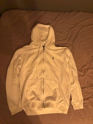 Vit hoodie från Polo Ralph Lauren - Säljer en vit hoodie från Polo Ralph Lauren med dragkedja framtill och klassisk huva med dragsko. Tröjan har två stora fickor fram och den ikoniska lilla loggan broderad på bröstet. Tillverkad i mjukt bomullsmaterial och har ribbade muddar vid ärmslut och nederkant.