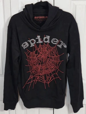 Sp5der Hoodie - Svart hoodie från Spider i 100% bomull med stor röd spindelnätsdesign och texten 'spider' i glittrande strass på bröstet. Hoodien har huva och ribbade muddar. Perfekt för dig som gillar edgy och unik streetstyle.