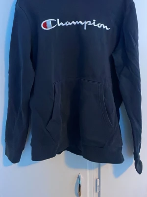 Mörkblå Champion hoodie med logga - Snygg mörkblå hoodie från Champion med stor broderad logga i vitt, rött och blått på bröst, samt klassisk känguruficka framtill. Mjuk bom drillskvalitet och huva. Perfekt för en avslappnad everywhere-look.