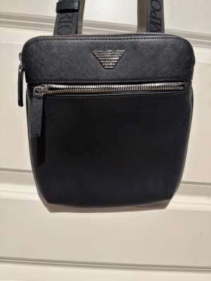 Svart axelväska från Emporio Armani - Snygg svart axelväska från Emporio Armani med silverfärgad logga framtill och matchande dragkedjor. Väskan har justerbart axelband med broderad logga och är tillverkad i ett tåligt, strukturerat material. Perfekt för att bära det viktigaste med stil.(ÄKTA) möts upp oxå i Sthlm 