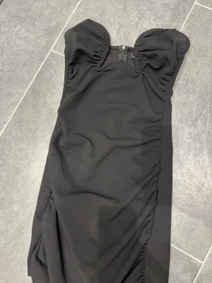 Svart axelbandslös kortklänning - Snygg svart axelbandslös kortklänning med rynkade detaljer och markerad byst. Klänningen har en figurnära passform och dragkedja bak. Perfekt för dig som vill ha en stilren och trendig look.