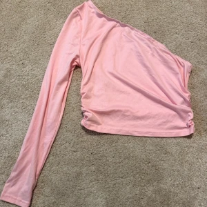 Rosa oneshoulder topp från SHEIN - Snygg rosa oneshoulder topp från SHEIN med lång ärm på ena sidan och rynkade detaljer i sidan. Tillverkad i mjukt och stretchigt material med en tight passform. Perfekt för dig som vill sticka ut lite extra!