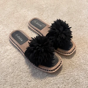 Svarta sandaler med blomdetalj Joia - Unika sandaler från Joia med stora svarta blomdetaljer på ovandelen. Sandalerna har fyrkantig tå, flätad beige sula och svart innersula med Joia-logga. Perfekta för dig som vill sticka ut med en cool och trendig look.