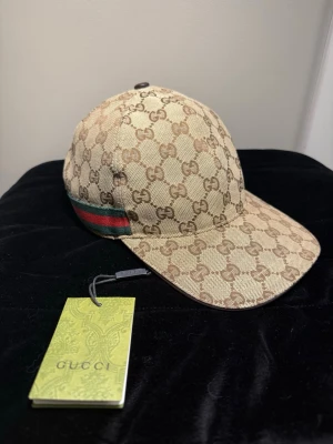 Gucci keps med GG-mönster - Snygg beige keps från Gucci med klassiskt GG-monogram över hela kepsen. Kepsen har en böjd skärm och en ikonisk grön och röd rand på sidan. Tillverkad i canvasmaterial som ger en exklusiv känsla och streetig look.