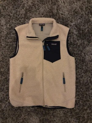 Fleeceväst från Patagonia, offwhite - Säljer en mysig offwhite fleeceväst från Patagonia med marinblå detaljer. Västen har dragkedja framtill, två sidofickor med blå zippar och en bröstficka i marinblått tyg. Perfekt för lager-på-lager och chill stil.