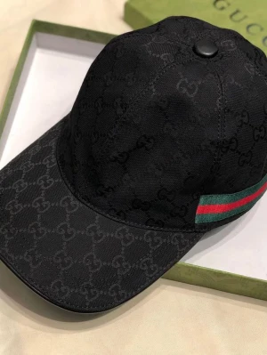 Svart Gucci keps med logomönster - Säljer en svart keps från Gucci med diskret GG-logomönster över hela kepsen. På sidan finns en klassisk grön och röd rand. Kepsen har böjd skärm och är tillverkad i canvasmaterial. Perfekt för dig som vill ha en exklusiv och trendig accessoar.