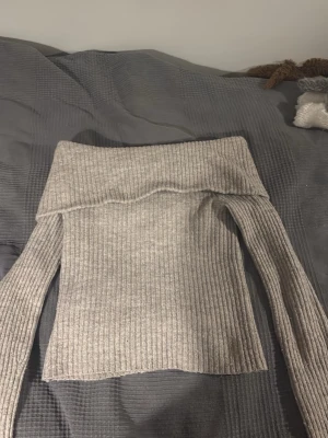 Ljusgrå ribbad offshoulder stickad tröja - Trendig ljusgrå stickad tröja med ribbad struktur och bred offshoulder-krage. Långärmad modell som sitter snyggt och passar perfekt till både jeans och kjol. Supermjuk och skön för kyligare dagar.