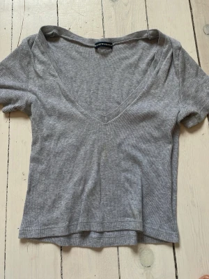 Grå ribbad cropped topp från Brandy Melville - Säljer en ljusgrå cropped ribbad topp från Brandy Melville med v-ringning och korta ärmar. Toppen har en croppad passform och är tillverkad i mjuk bomullsmix som känns skön mot huden. Perfekt att styla med jeans eller kjol.