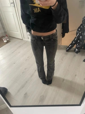 Svarta low waist bootcut jeans - Säljer ett par svarta low waist jeans med bootcut passform. Jeansen har slitna detaljer, vita sömmar och dekorativa knappar på bakfickorna. Perfekta för en trendig och avslappnad stil. (Endast andvända 3 gånger)