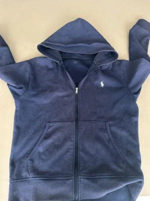 Mörkblå hoodie från Polo Ralph Lauren - Mörkblå hoodie med dragkedja från Polo Ralph Lauren. Klassisk design med huva, två fickor framtill och den ikoniska vita logotypen broderad på bröstet. Tillverkad i mjukt bomullsmaterial som känns skönt mot huden. Perfekt för en avslappnad och stilren look.