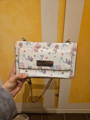 Vit axelväska med sjöjungfrumotiv - ❗️Skriv innan köp❗️Säljer en vit axelväska med färgglada sjöjungfrur, sjöstjärnor och havsmotiv. Väskan har en silverfärgad kedjerem och magnetlås. Materialet är konstläder med struktur och väskan har en rektangulär form. Inuti är väskan missfärgad pga en kvarliggande godis som luktat skit❤️, men annars är väskan i fint skick.