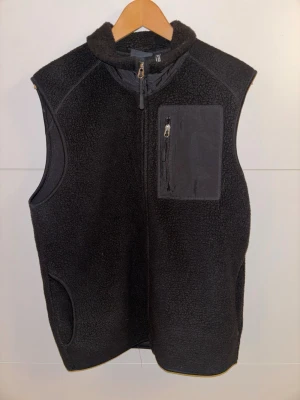 Svart fleeceväst från H&M - Svart fleeceväst från H&M med hög krage och hel dragkedja framtill. Västen har en praktisk bröstficka med dragkedja och detaljer i nylon. Perfekt för lager-på-lager och riktigt skön att bära under kyliga dagar.