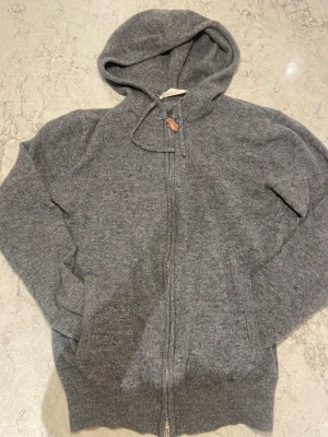 Grå cashmere hoodie från Sayless - Mjuk och lyxig hoodie i grått från Sayless, tillverkad i cashmere. Hoodien har dragkedja framtill, huva med dragsko och ribbade muddar vid ärmslut och nederkant. Perfekt för dig som gillar stilrena och bekväma plagg.