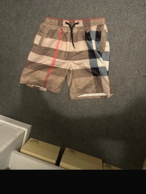 Beige rutiga shorts med snörning - Snygga beige shorts från Burberry med klassiskt rutigt mönster i brunt, svart, vitt och rött. Shortsen har elastisk midja med svart snörning och är perfekta för sommaren. Lätta och bekväma med en modern passform.