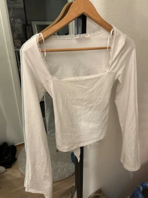 Vit långärmad omlottopp med spets - Snygg vit långärmad topp med omlottdetalj och knyt i ryggen. Toppen har fyrkantig ringning och spetskant runt halsen för en söt touch. Helt ny och aldrig använd!