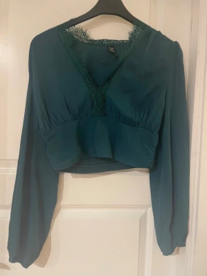 Grön croppad blus med spetsdetalj - Snygg mörkgrön croppad blus med v-ringning och spetsdetaljer vid halsen. Blusen har långa, luftiga ärmar och smockad midja för en bekväm passform. Perfekt att styla med höga jeans eller kjol.